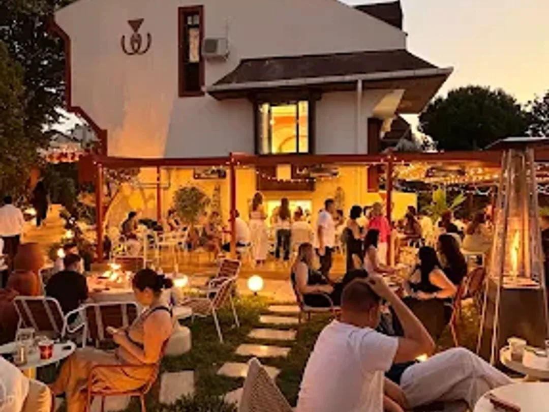Akşamüstü Ruum: aydınlatılmış villa cephesi, bahçedeki dolu masalar ve turuncu pergola